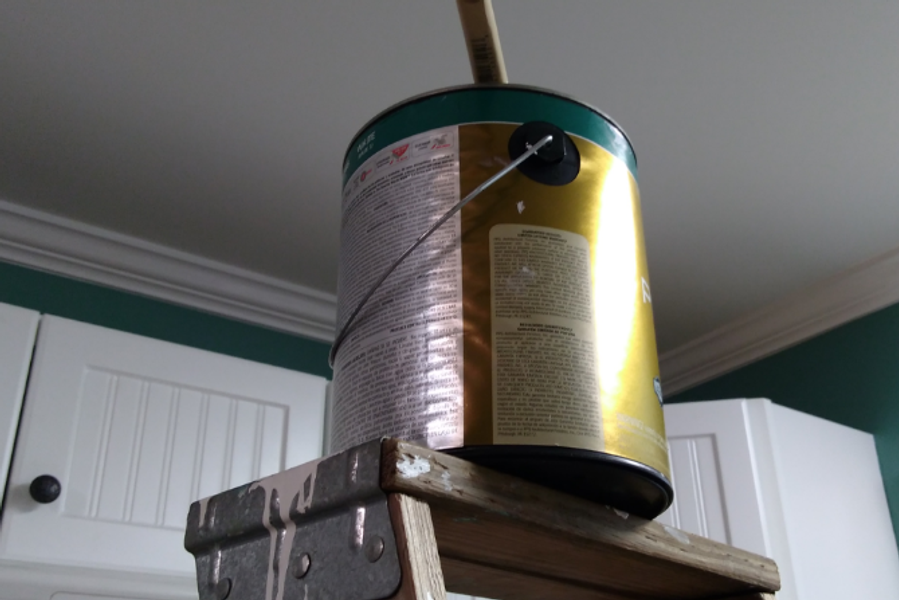 A gallon paint can with a brush resting across its rim sits on top of a wooden stepladder in a kitchen with green walls and white cabinets.