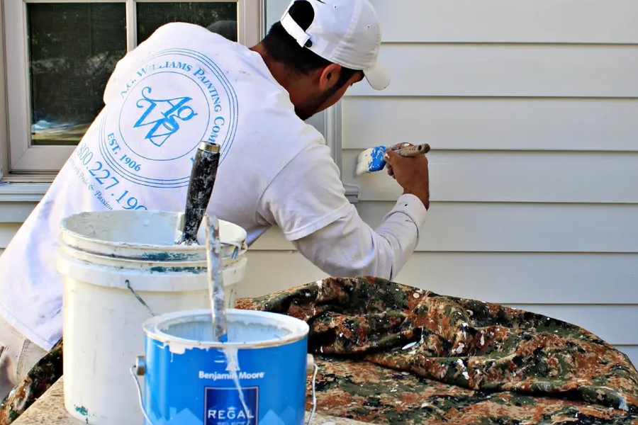 A painter in a white A.G. Williams Painting Company shirt marked "Est. 1906" brushes light gray paint onto house siding beside a Benjamin Moore Regal can and a white bucket.