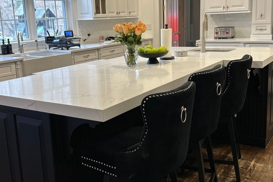 Renovated kitchen with a large black-painted island topped in white marble-look quartz, three black tufted barstools, white perimeter cabinets, and orange roses in a glass vase.