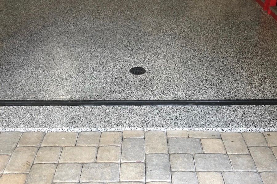 Speckled gray epoxy-coated garage floor with a small drain, meeting a paver-stone driveway apron at the garage door threshold.
