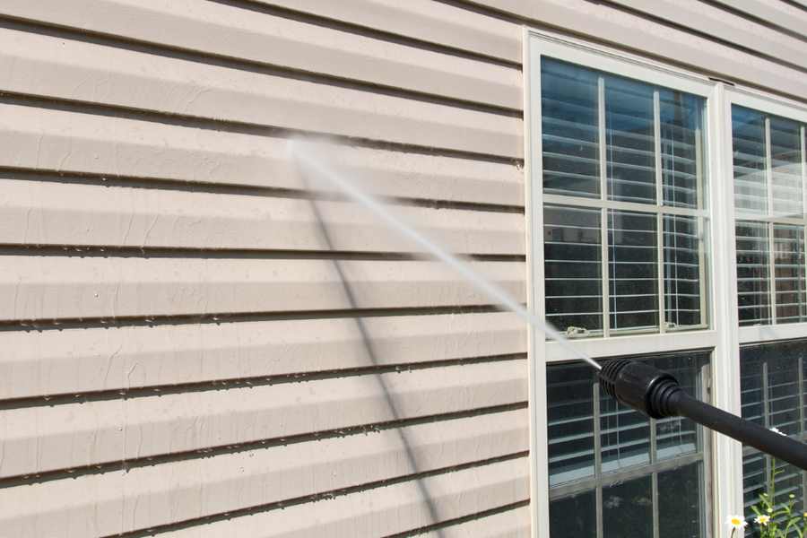 A pressure-washer wand sprays a jet of water across beige horizontal siding beside white double-hung windows on a home's exterior.