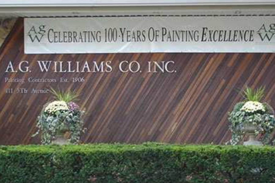 Exterior sign for A.G. Williams Co. Inc., Painting Contractors Est. 1906, on a wood-paneled building, with a banner above reading "Celebrating 100 Years of Painting Excellence" behind hedges and potted flowers.