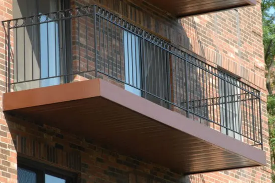 Balcony with a black wrought-iron railing and copper-toned deck projecting from a red brick apartment building, with sliding glass doors and trees in the background.