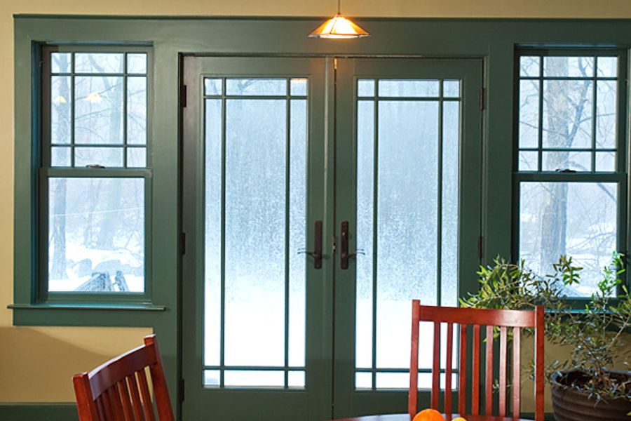 Dining room with dark green-trimmed French doors and windows overlooking a snowy yard, framed by yellow walls, wooden chairs, and a potted plant.