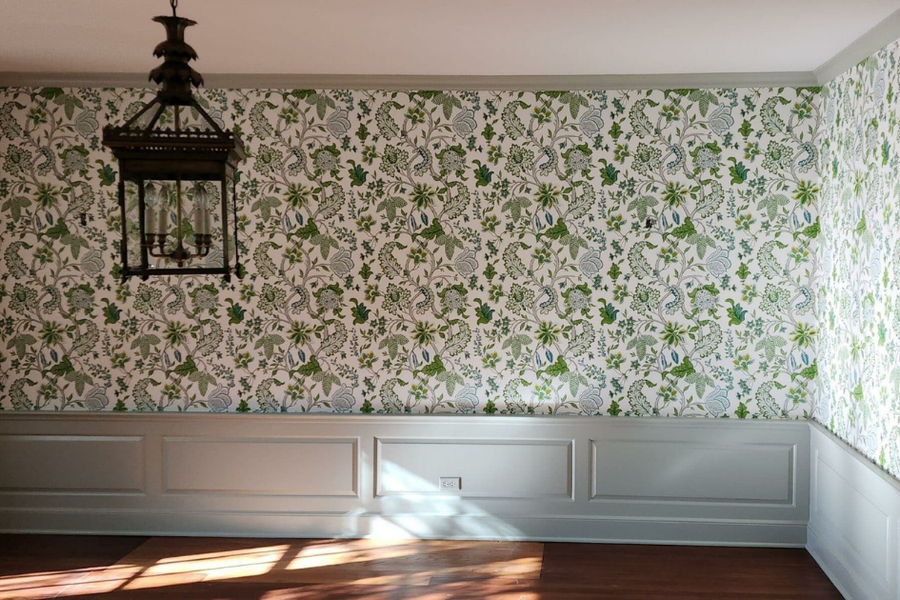 Dining room with green-and-white botanical wallpaper above gray painted wainscoting, hardwood floors, and a bronze lantern-style pendant light hanging from the ceiling.