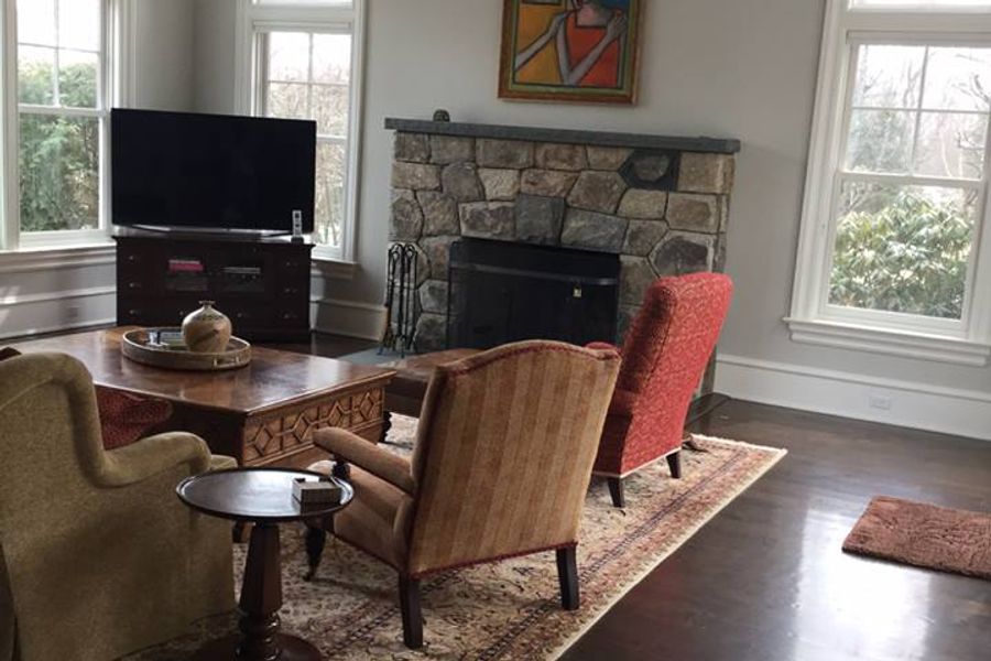 Living room with light gray walls, a stone fireplace, upholstered chairs on a patterned rug, and a white poodle resting on the dark hardwood floor.