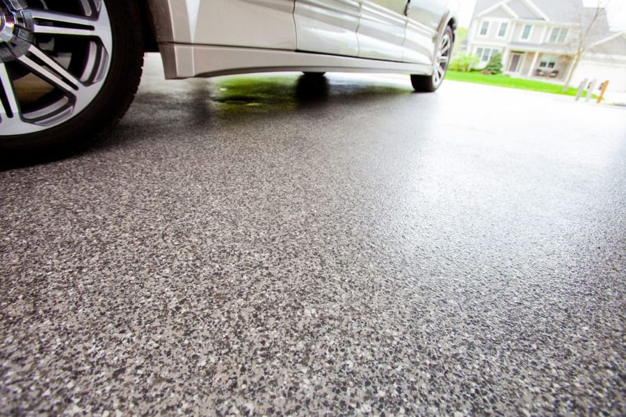 Close-up of a gray speckled epoxy-coated garage floor beneath a parked silver car, with a suburban house and lawn visible through the open garage door.