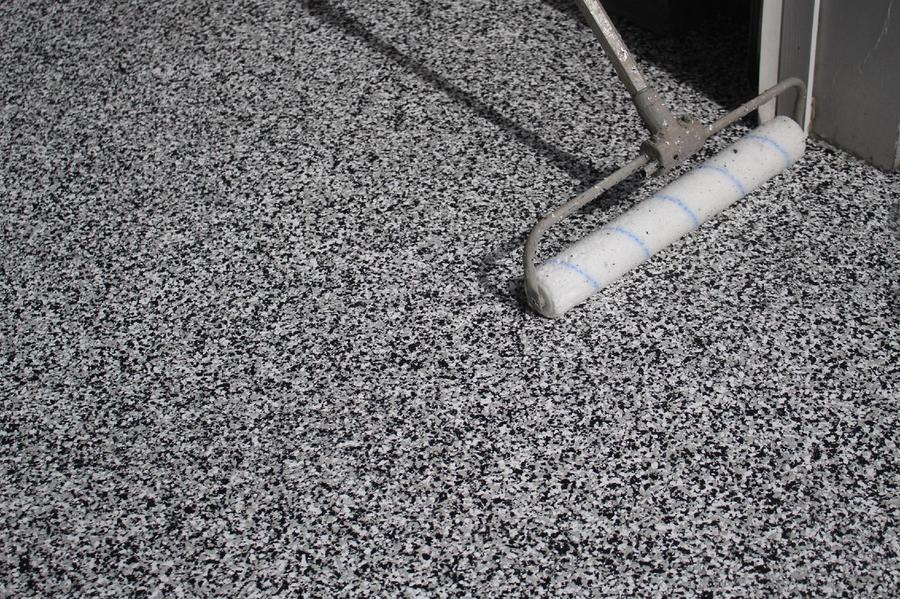 A long-handled roller applies clear coating over a freshly installed black, white, and gray flake epoxy floor near a doorway.