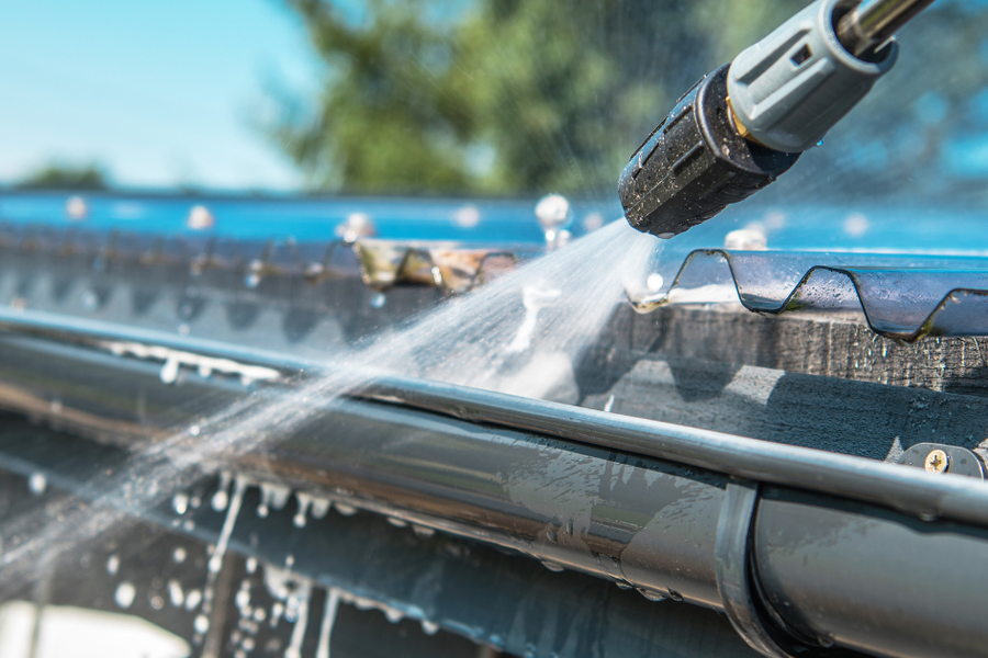 A pressure-washer nozzle sprays water into a metal roof gutter on a sunny day, with droplets splashing and trees in the background.