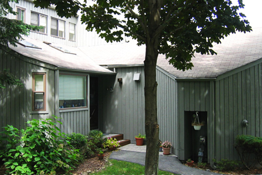 Condominium exterior with gray-green vertical wood siding, a shaded front entry, potted flowers along the walkway, and a large tree in the foreground.