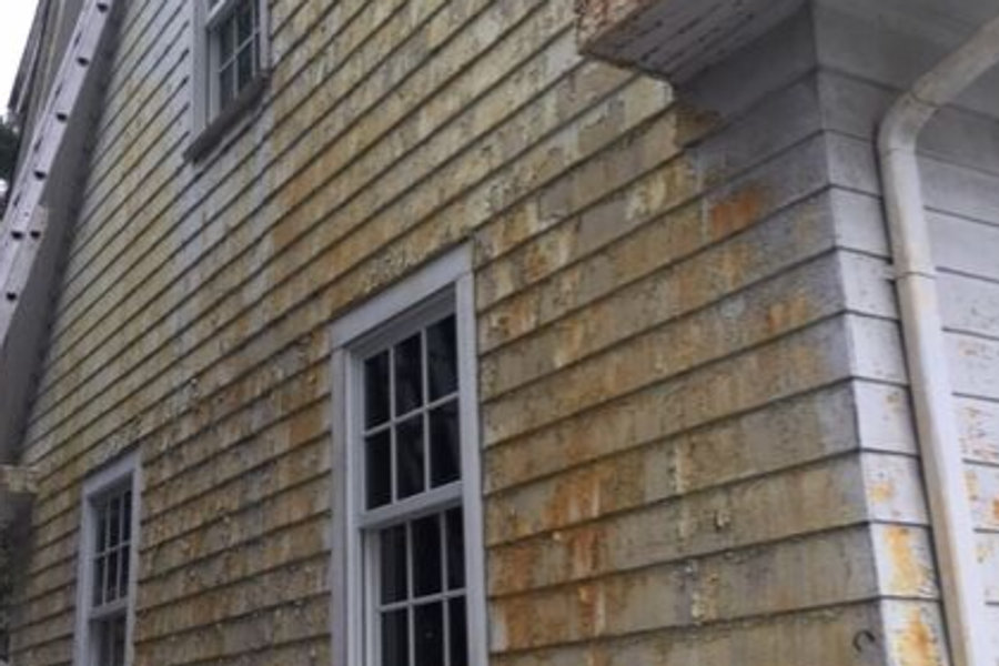 Side of a shingled house with severely failing exterior paint, showing widespread peeling, rust-colored staining, and bare weathered wood around white-trimmed windows.