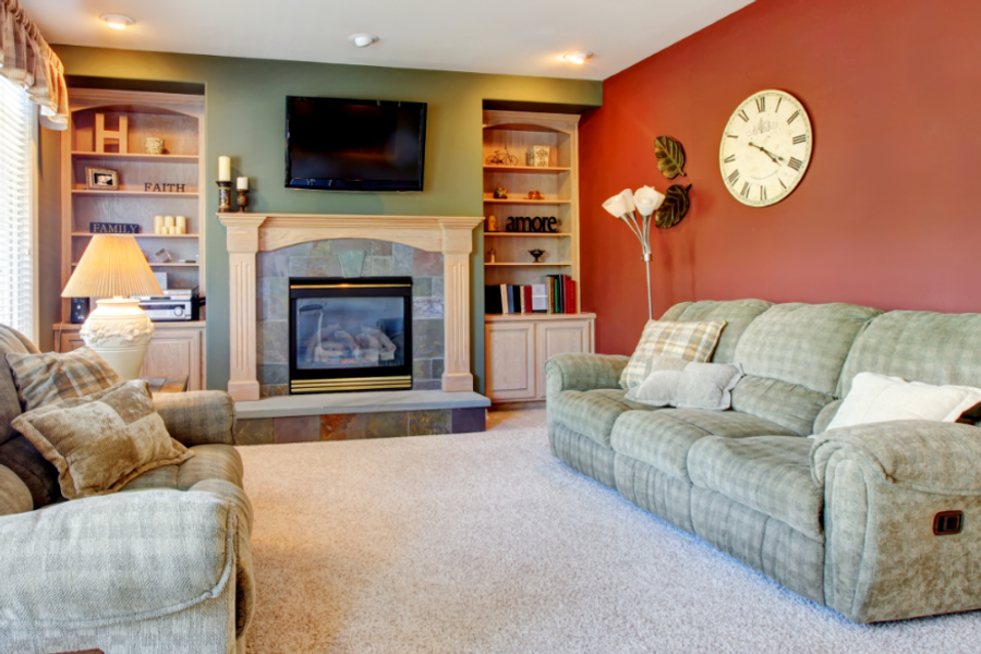 A living room with a deep red accent wall and sage green fireplace wall, featuring plush sofas, built-in shelves, a wall clock, and a stone-surround fireplace.