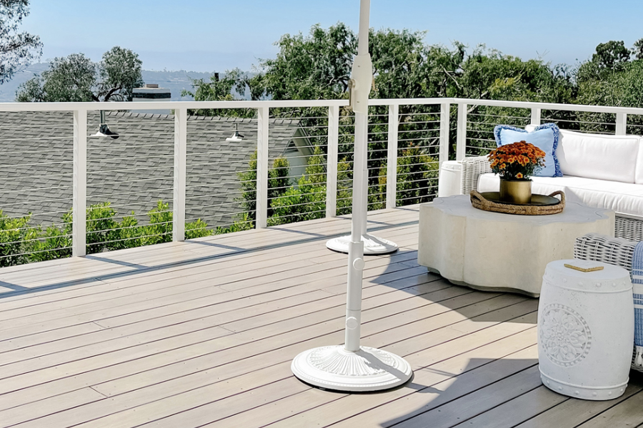 Sunlit rooftop deck with gray-stained boards, white cable railing, a wicker sectional with blue pillows, and treetops in the distance.