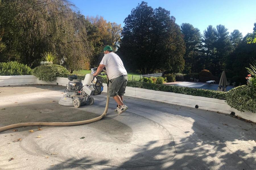A worker in a white shirt and green cap operates a concrete grinder with a dust-extraction hose on an outdoor patio, with a pool and trees behind a low hedge wall.