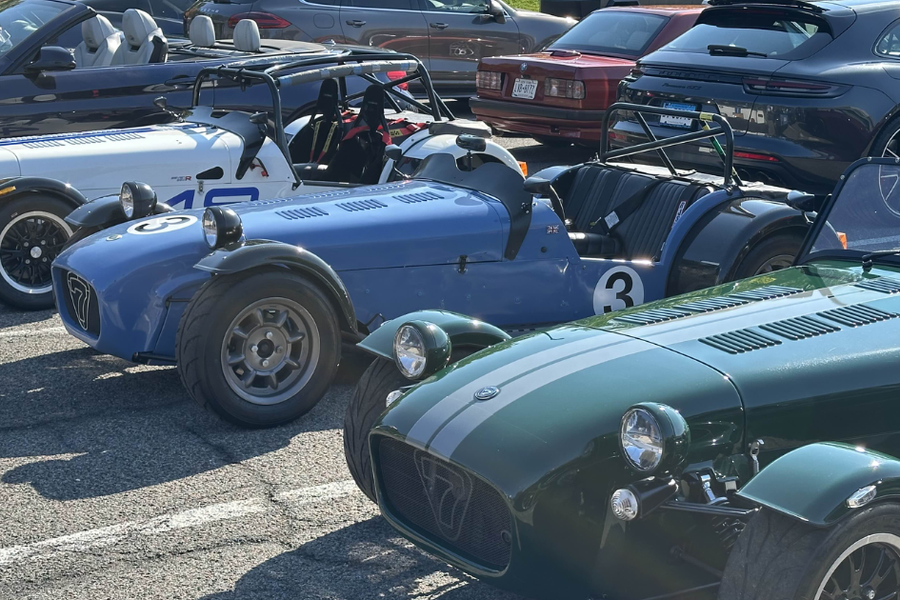 Blue, white, and green Caterham-style roadsters with racing numbers parked in a row at a sunny car meet, with other vehicles in the background.