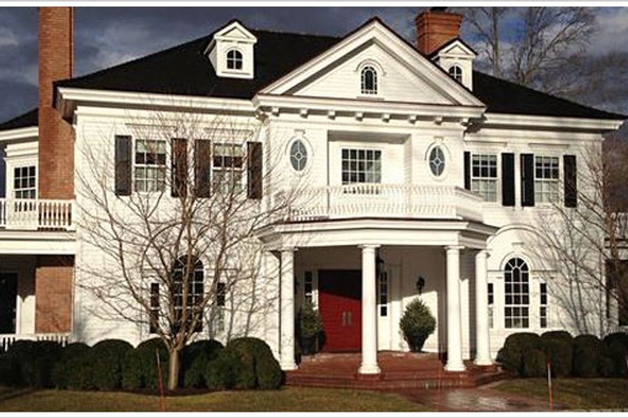 Large white colonial house with black shutters, columned front portico, red double doors, brick chimneys, and trimmed hedges under a cloudy sky.