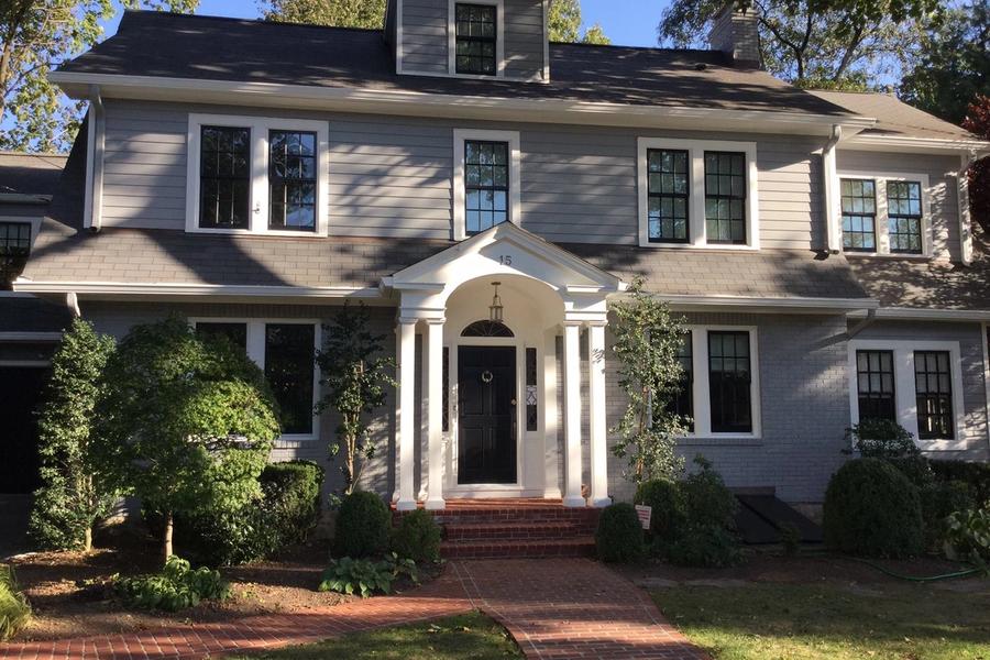 Two-story gray colonial home with white trim, a columned front portico, black front door, and a red brick walkway lined with shrubs and landscaping.