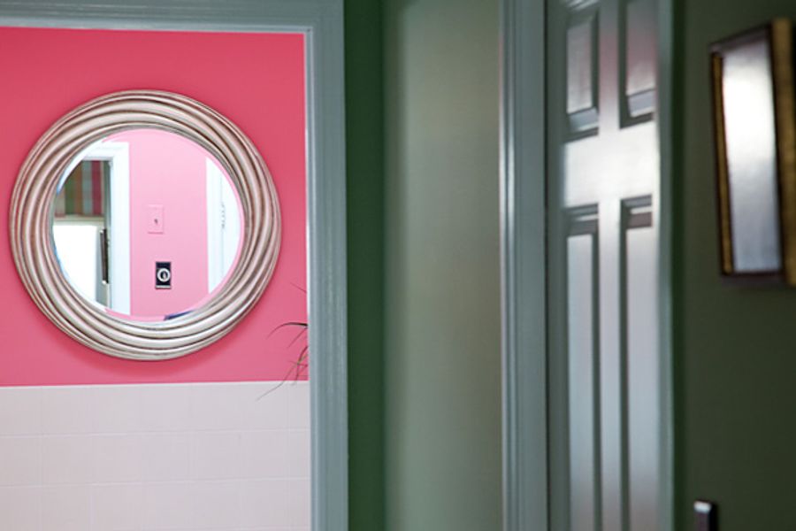 View through a doorway into a bathroom with bright pink walls, white tile wainscoting, and a round silver-framed mirror, seen from a green-painted hallway.