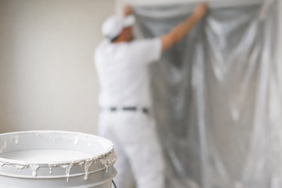 Open bucket of white paint in the foreground while a painter in white clothing and cap hangs plastic sheeting over a wall behind it.