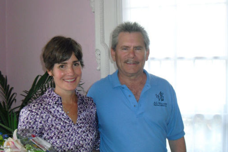 A woman in a patterned blouse and a man in a light blue A.G. Williams polo shirt stand smiling indoors beside a flower arrangement and a curtained window.