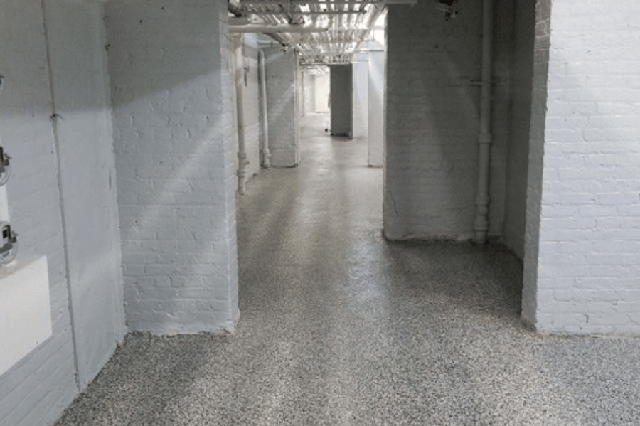 Basement corridor with freshly painted white brick walls and exposed ceiling pipes, finished with a gray speckled epoxy flake floor coating.