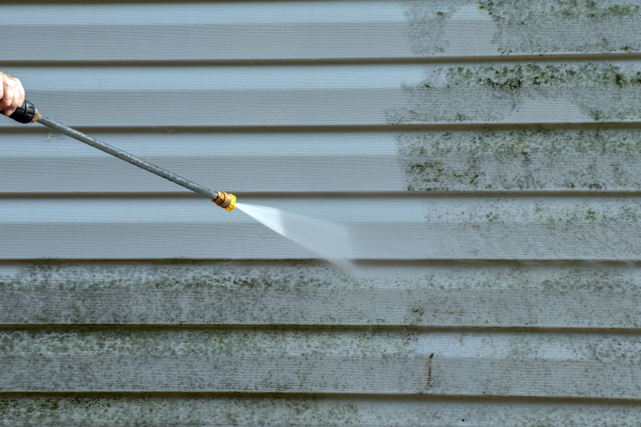 A hand aims a pressure-washer wand at mildew-stained vinyl siding, spraying a clean white stripe across the green-streaked surface.