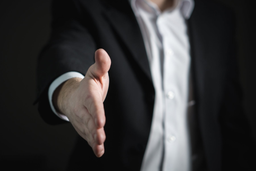 A person in a dark suit and white shirt extends an open hand toward the camera for a handshake against a dark background.