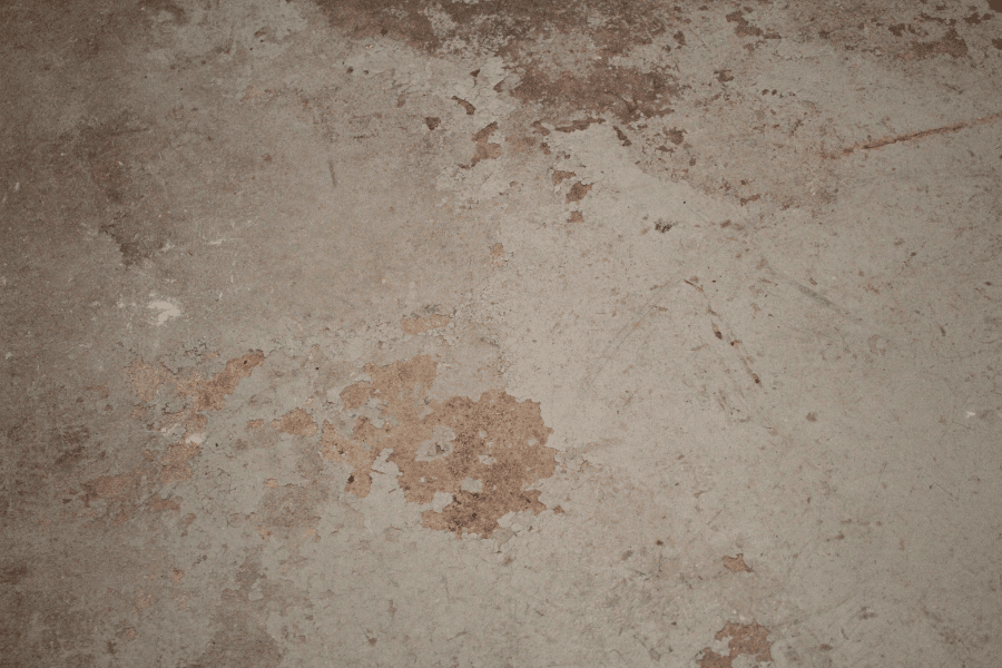 Close-up of a worn concrete floor surface with brown staining, scratches, and patches of flaking material across the gray finish.