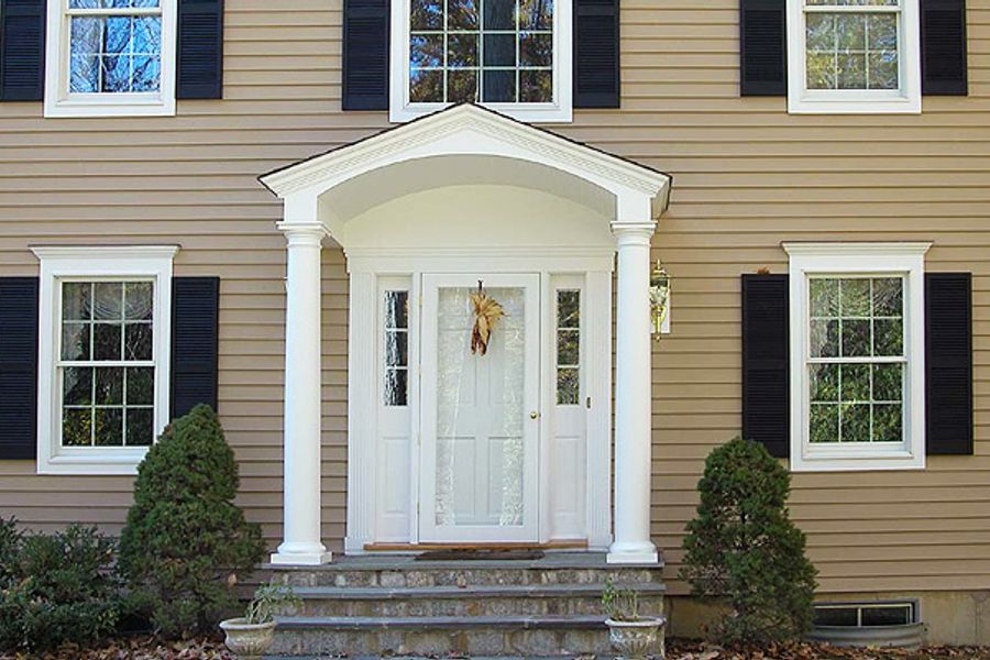 Front entry of a tan clapboard house with a white columned portico, black shutters, white six-panel door with sidelights, and stone steps flanked by shrubs.