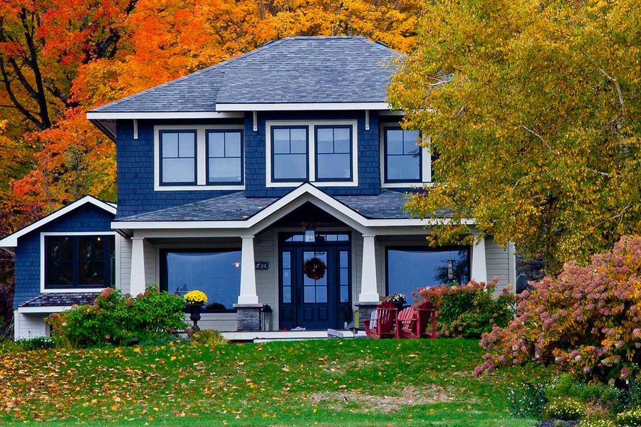 Two-story blue craftsman-style house with a covered front porch, surrounded by trees in bright orange and yellow fall foliage and a leaf-scattered lawn.