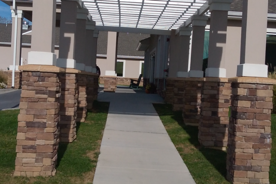 Covered walkway at a senior living facility, with a white pergola supported by stacked-stone and white columns leading along a concrete path toward the entrance.