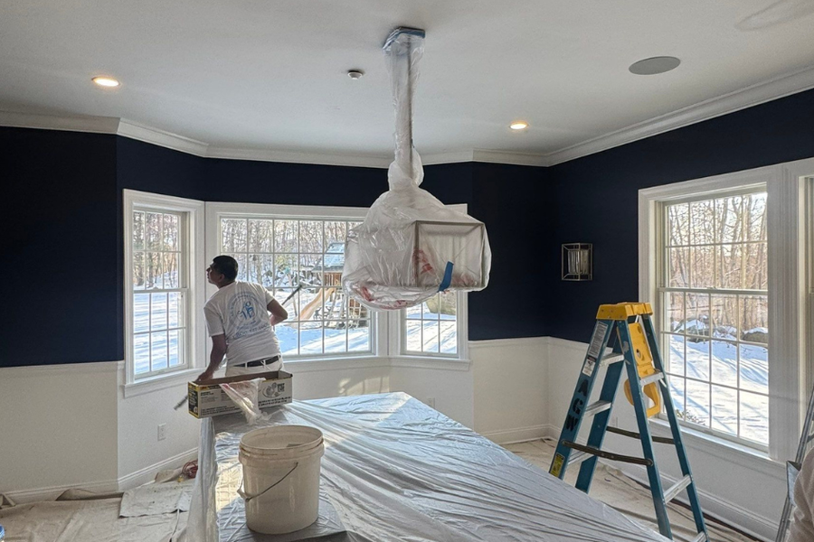 A painter in a white shirt works near bay windows in a navy-blue room with white wainscoting, with a plastic-wrapped chandelier, covered furniture, and ladders during ceiling prep.
