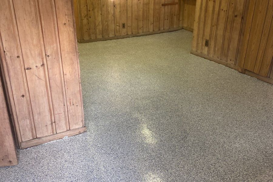 Freshly coated basement floor with a gray flake epoxy finish in a room with knotty pine wood-paneled walls and exposed ceiling joists.