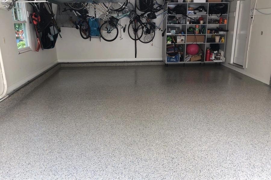 Garage interior with a freshly coated gray speckled epoxy floor, bicycles hanging from ceiling racks, and shelving units of household gear along the back wall.