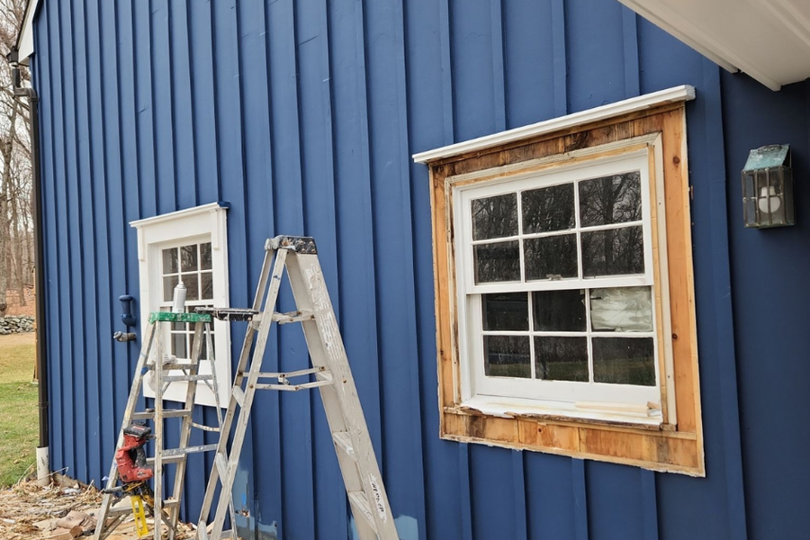 A blue board-and-batten house exterior with an aluminum stepladder set up beside a window whose white sash sits in freshly stripped bare-wood trim awaiting paint.
