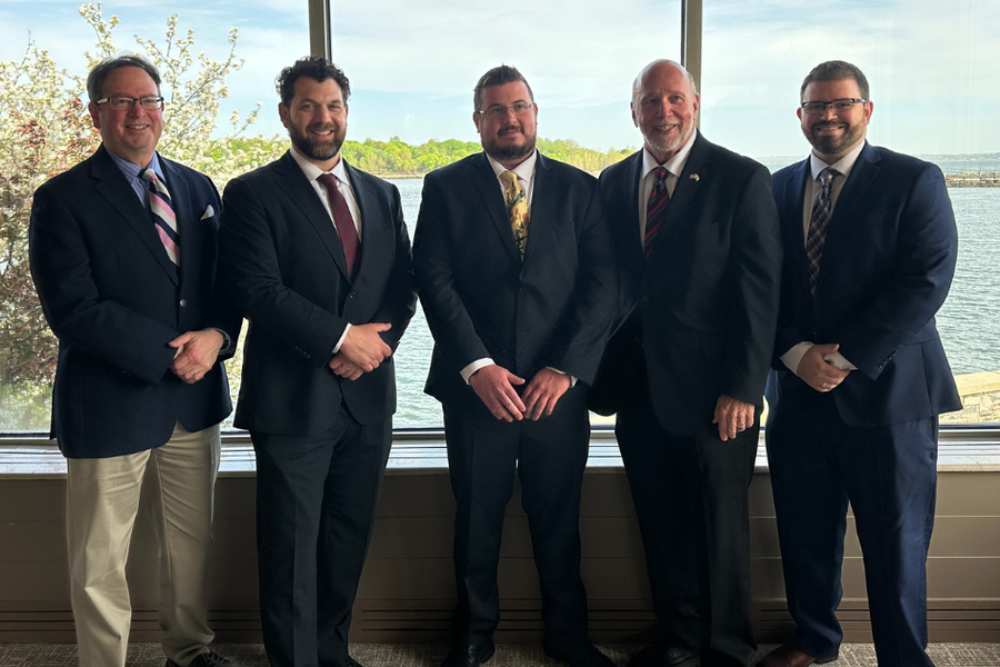 Five men in suits and ties stand together in front of a large window overlooking a waterfront with trees and a stone breakwater.