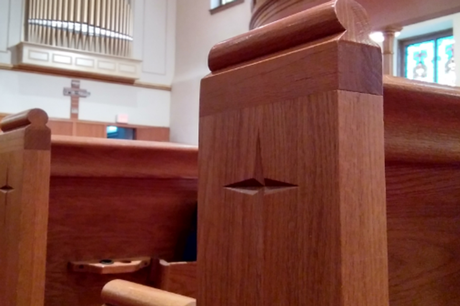 Wooden church pews with a carved star detail on a pew post, with a pipe organ, cross, arched window, and curved balcony in the background.