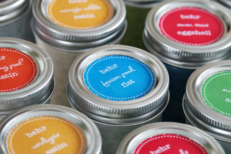 Small mason jars of leftover paint with colorful round Behr labels on the lids noting color names and finishes such as eggshell, satin, flat, and semi gloss.
