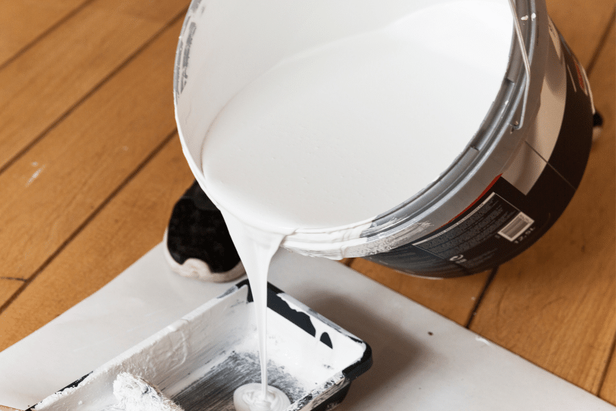 White paint pours from a large can into a roller tray set on protective paper over a wood floor, with a person's sneaker visible behind.