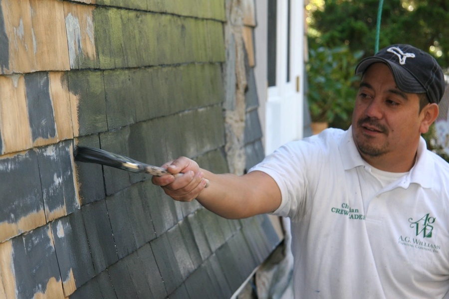 A painter in a white A.G. Williams crew leader shirt and black cap brushes gray paint onto weathered cedar shingle siding on a home exterior.