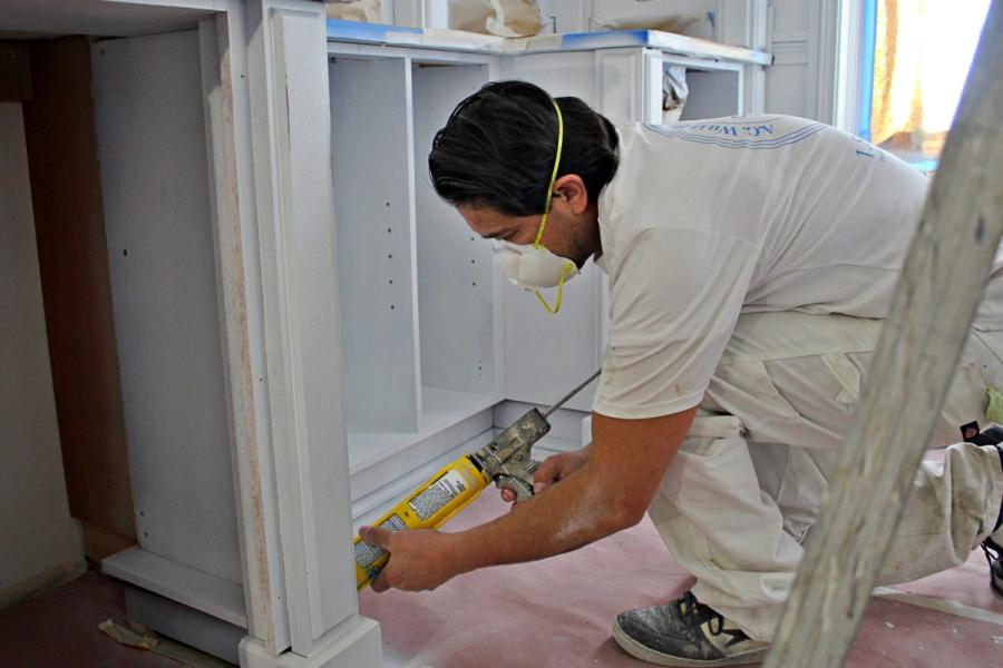 A painter in a white A.G. Williams shirt and dust mask kneels to apply caulk to white cabinet trim in a kitchen protected with rosy floor paper.