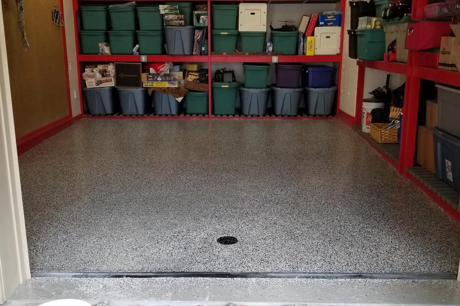 Garage with a freshly coated speckled gray epoxy floor, viewed from the open door, with red shelving units holding labeled storage bins along the back wall.