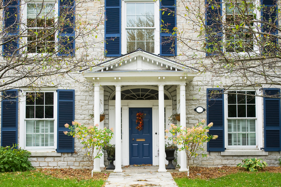 Stone colonial home with navy blue shutters and front door, a white columned portico, and hydrangeas flanking the entrance in autumn.