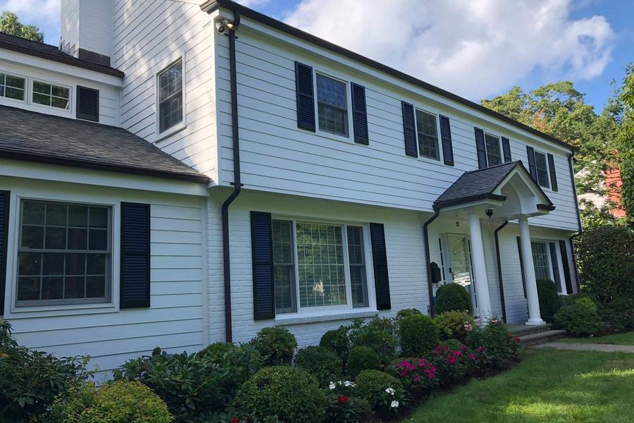 Two-story white colonial home with black shutters, a columned front portico, and freshly painted siding above tidy landscaping with flowering shrubs.