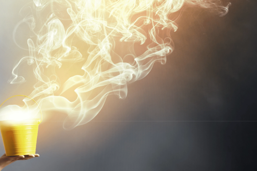 A hand holds up a small yellow paint bucket releasing swirls of glowing yellow vapor against a dark gray background.