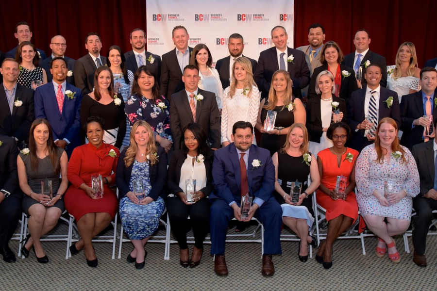 A large group of award recipients in business attire, many holding glass trophies, pose in rows before a backdrop with repeating BCW Business Council of Westchester logos.