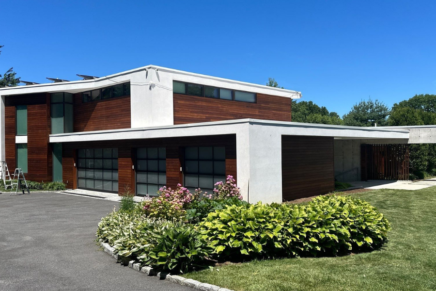 Modern two-story home with white stucco and dark wood siding under a clear blue sky, with ladders set up beside it and landscaped shrubs in front.