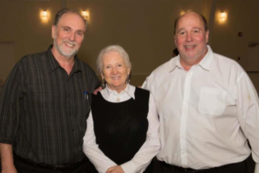 Two men in collared shirts stand smiling with an older woman in a black vest between them at an indoor event with wall sconces in the background.