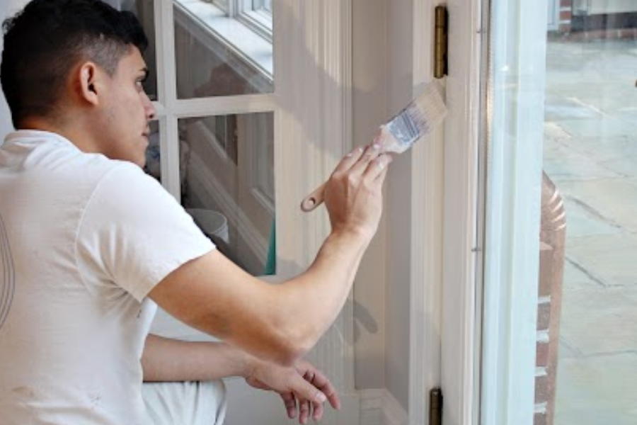 A painter in a white A.G. Williams Painting Company shirt brushes white paint onto interior French door trim beside a bluestone patio.