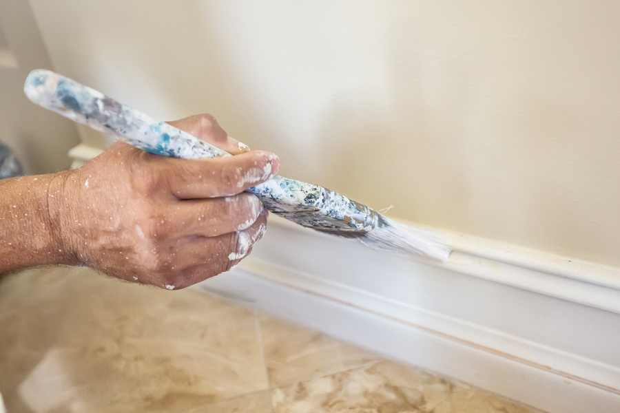 A paint-speckled hand guides a brush along white baseboard trim where it meets a beige wall above a polished tile floor.